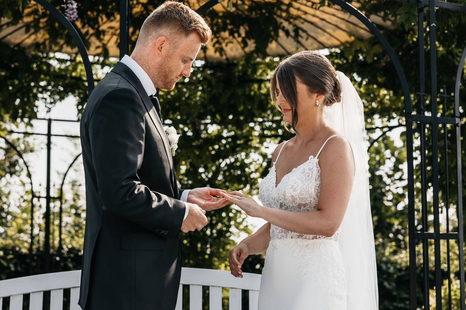 Groom places wedding ring on bride's finger beneath lush garden arbor during ceremony.