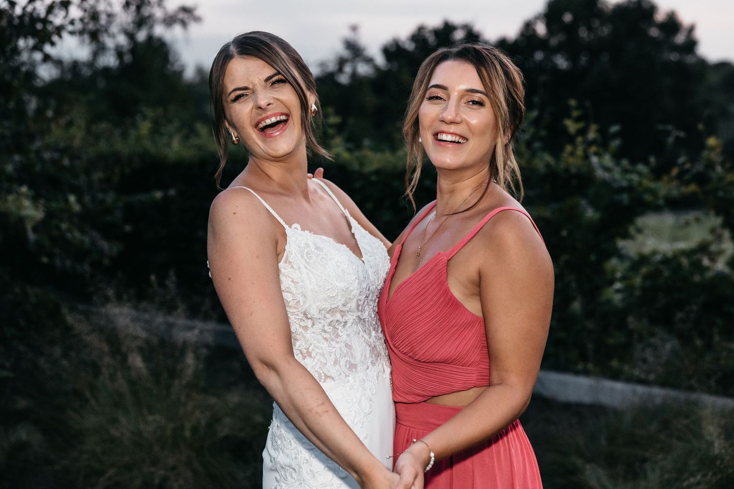 Laughing bride and bridesmaid in pink dress posing together at outdoor wedding celebration.