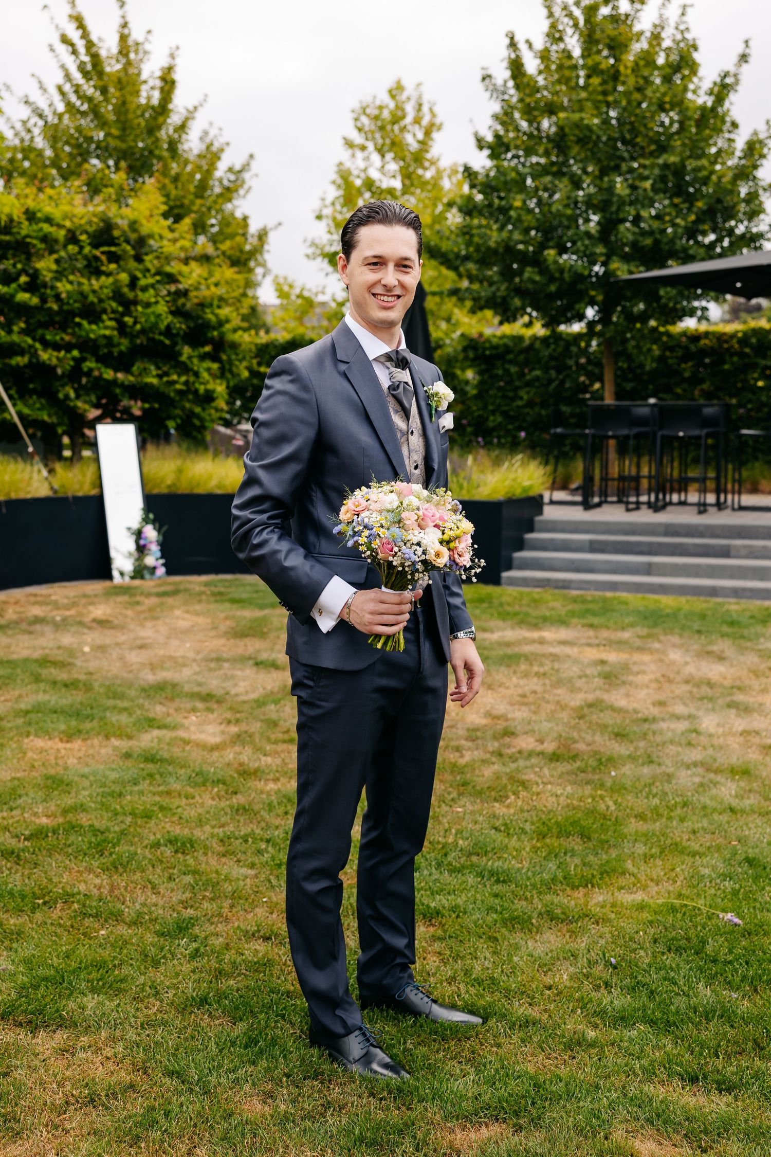 Smiling groom in navy suit holding colorful bridal bouquet standing on green lawn outdoors.