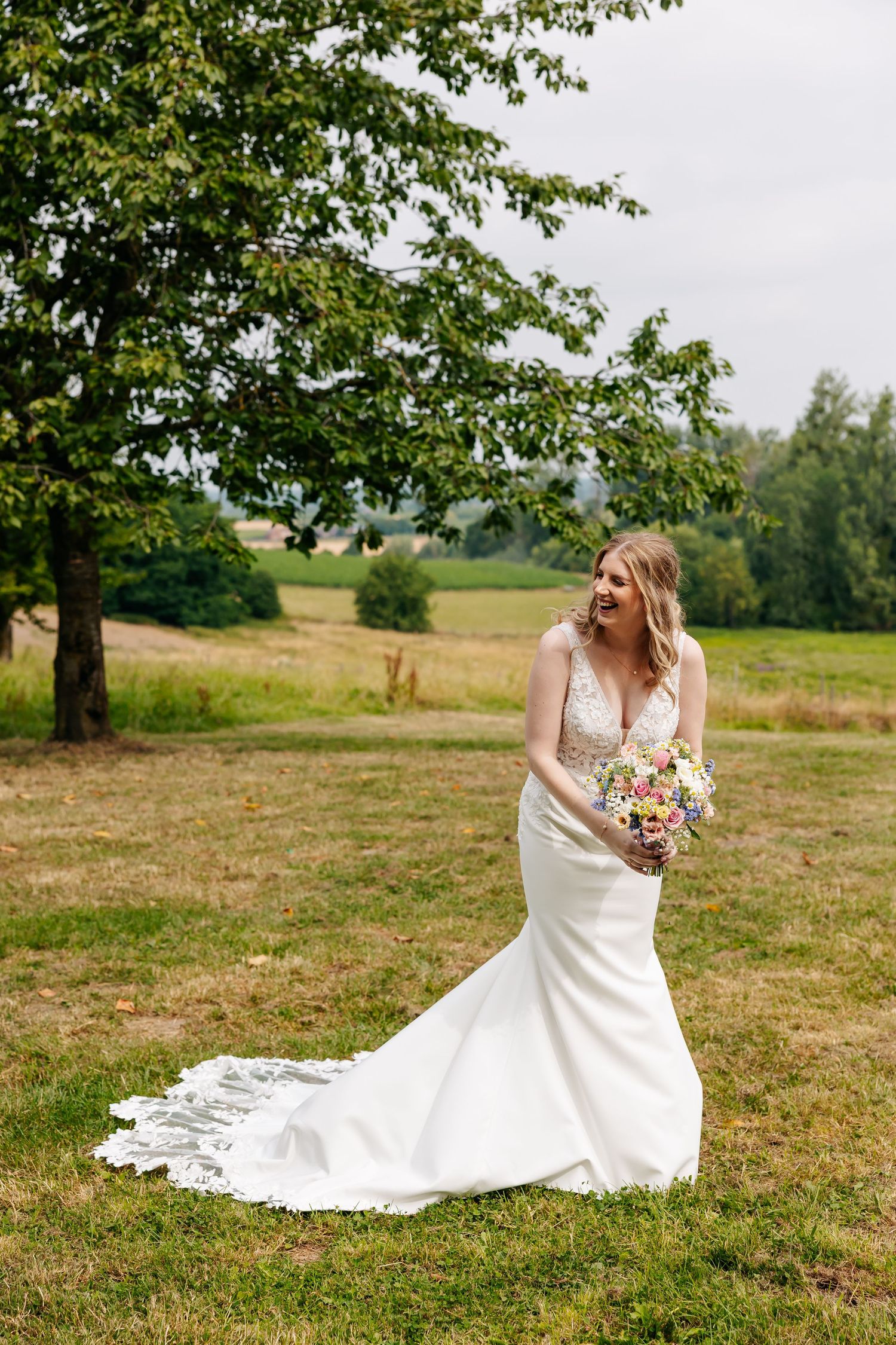 Bride in a fitted lace wedding dress laughing joyfully while holding a floral bouquet.