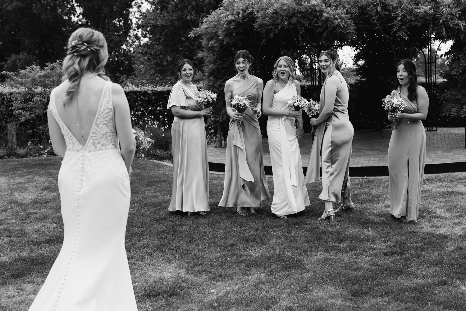 Black and white photo of bride facing excited bridesmaids holding bouquets on wedding day.