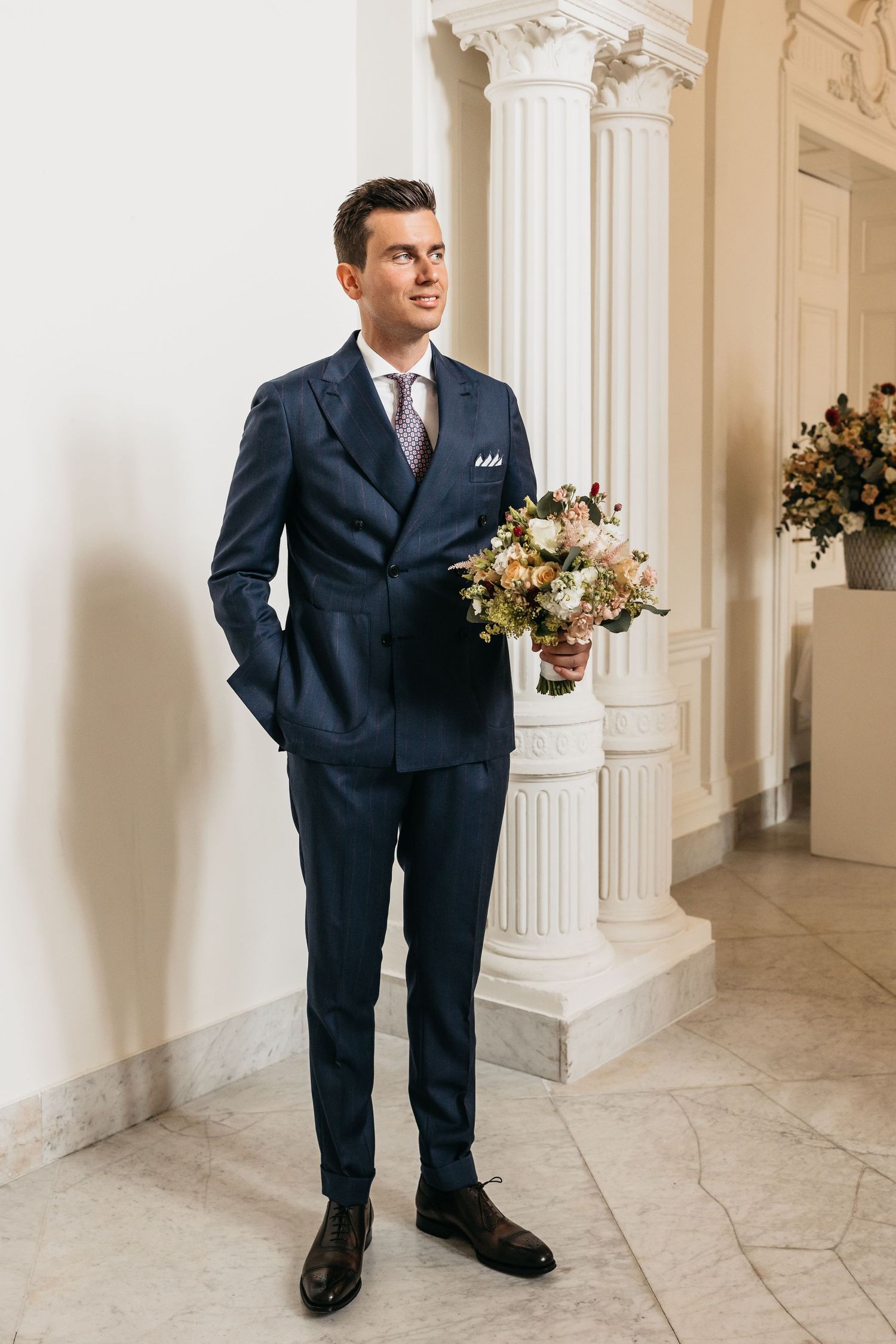 Dapper groom in navy pinstripe suit holding bridal bouquet beside white decorative columns.