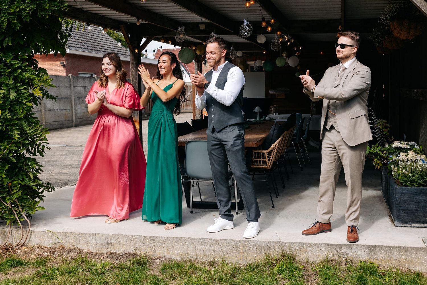 Wedding party members clap and cheer outdoors at a garden reception venue on a sunny day.