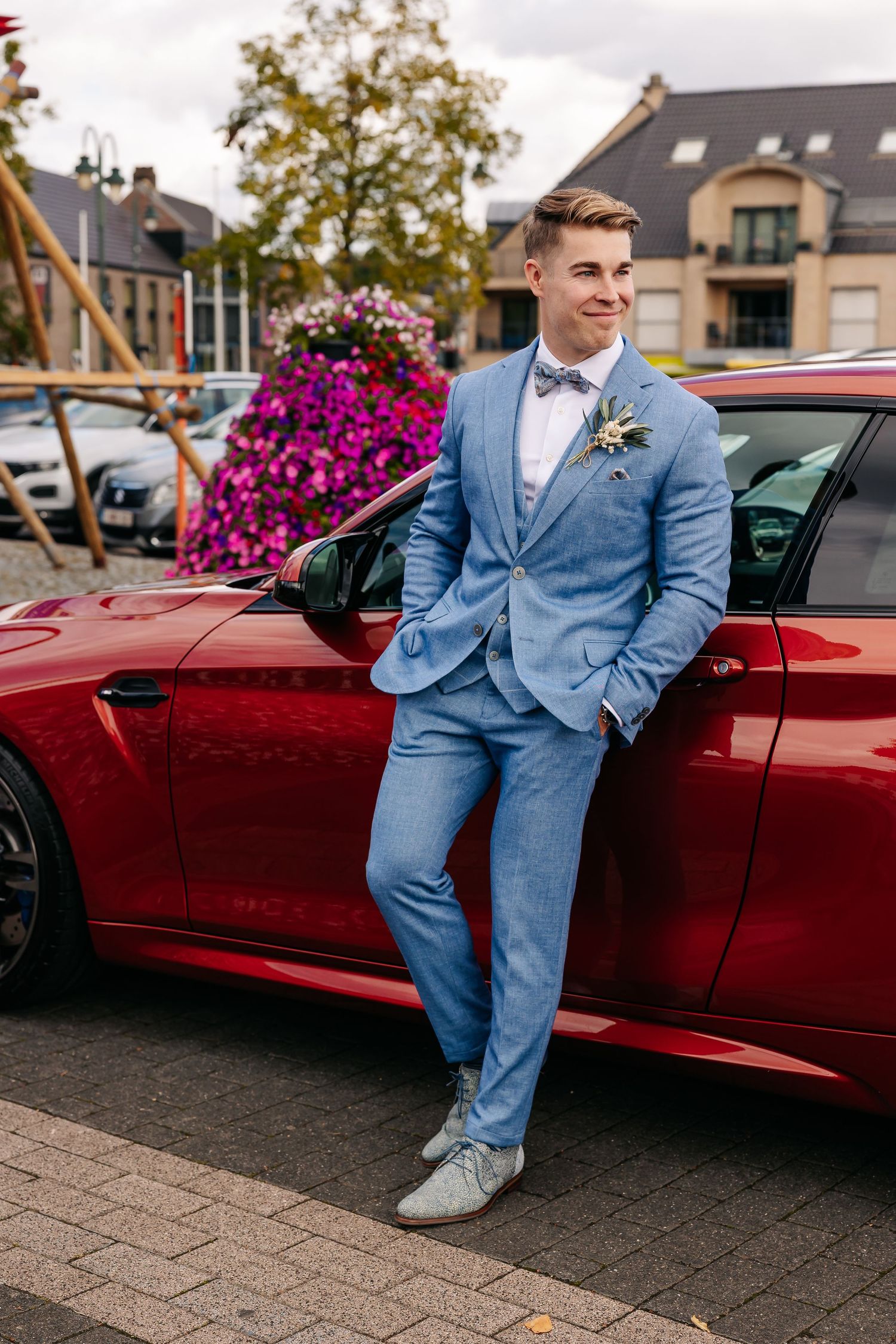 Stylish groom in blue suit leans against a red sports car at an outdoor wedding venue.