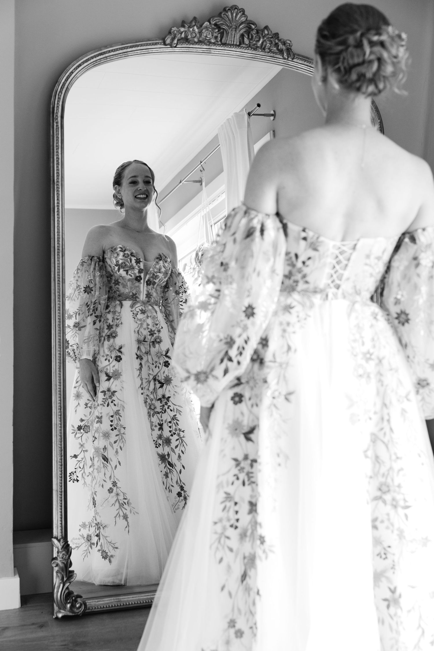 Bride in floral wedding gown smiling at her reflection in an ornate gold mirror, black and white photo.
