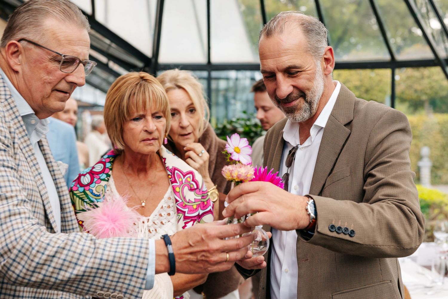 Group of wedding guests admiring pink flower in glass vase at outdoor celebration.