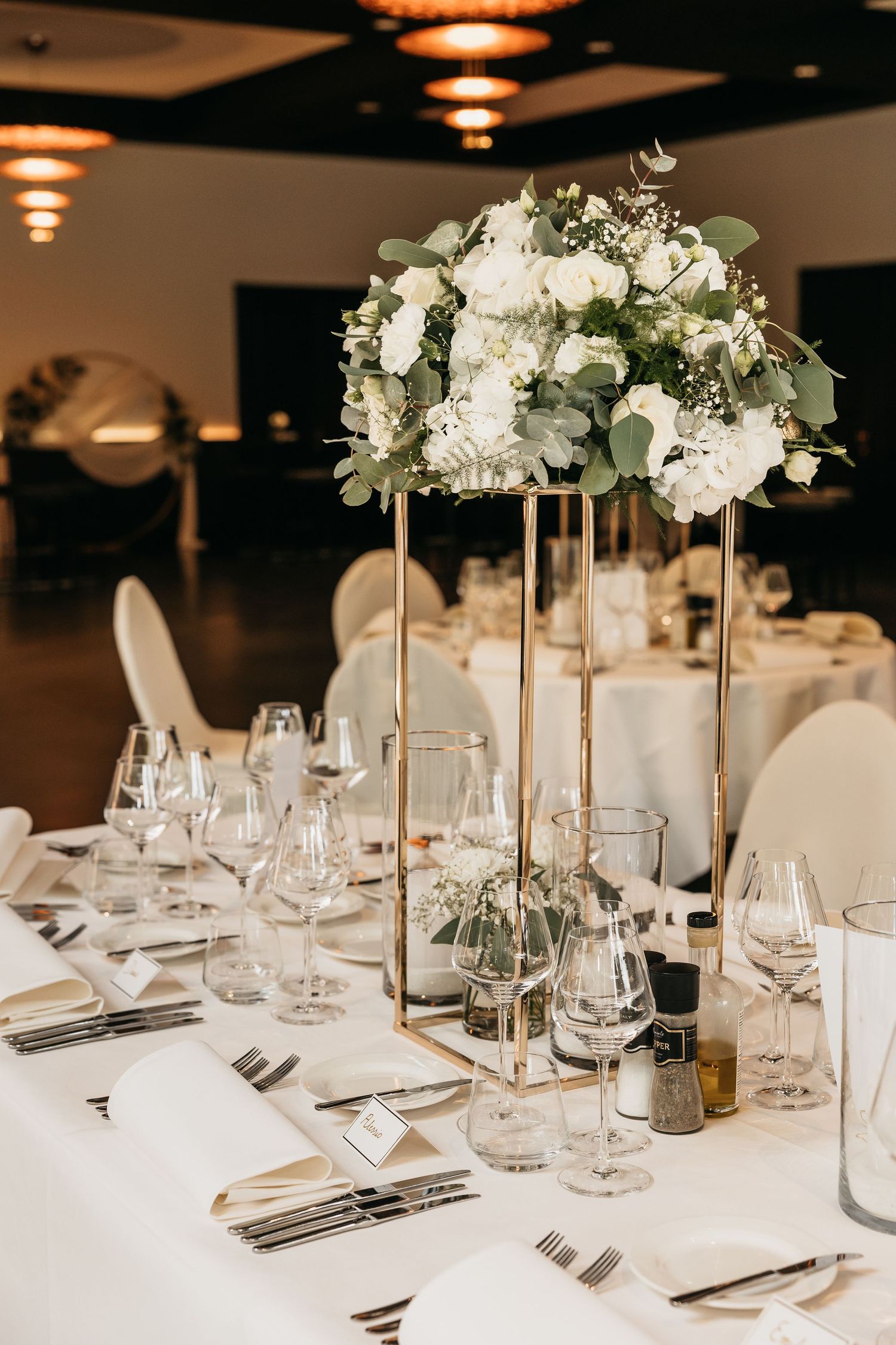 Tall gold geometric floral stand with white roses and eucalyptus centerpiece on a long wedding banquet table.