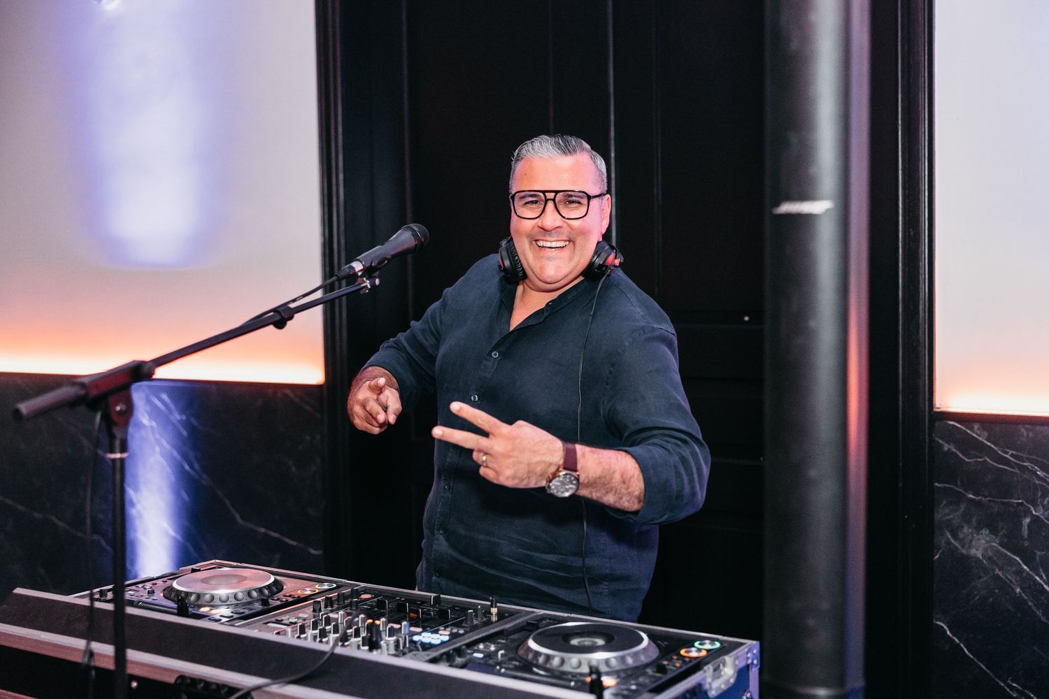 Smiling DJ giving peace sign behind turntables at wedding reception in modern venue.