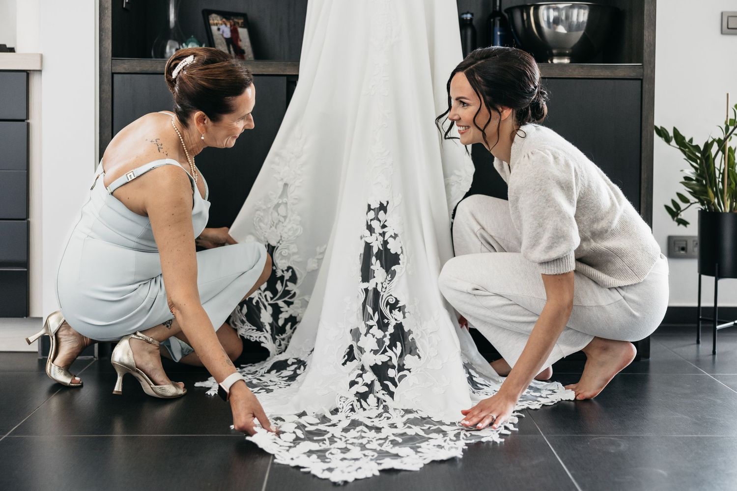 Two women crouching together admiring intricate lace wedding dress train on dark floor.