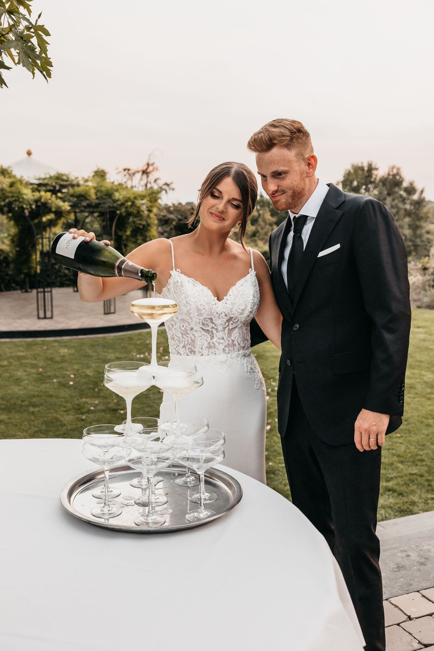 Bride and groom in elegant attire pouring champagne tower at outdoor wedding reception.