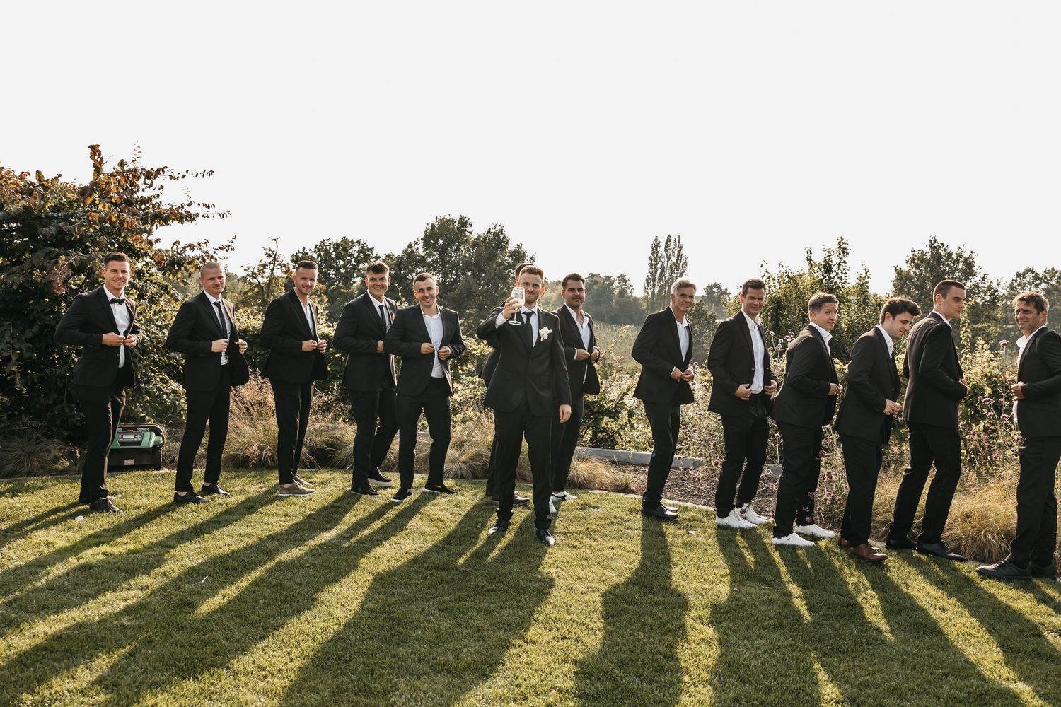 Groom and groomsmen in black suits laugh together on a sunny green lawn at a wedding.