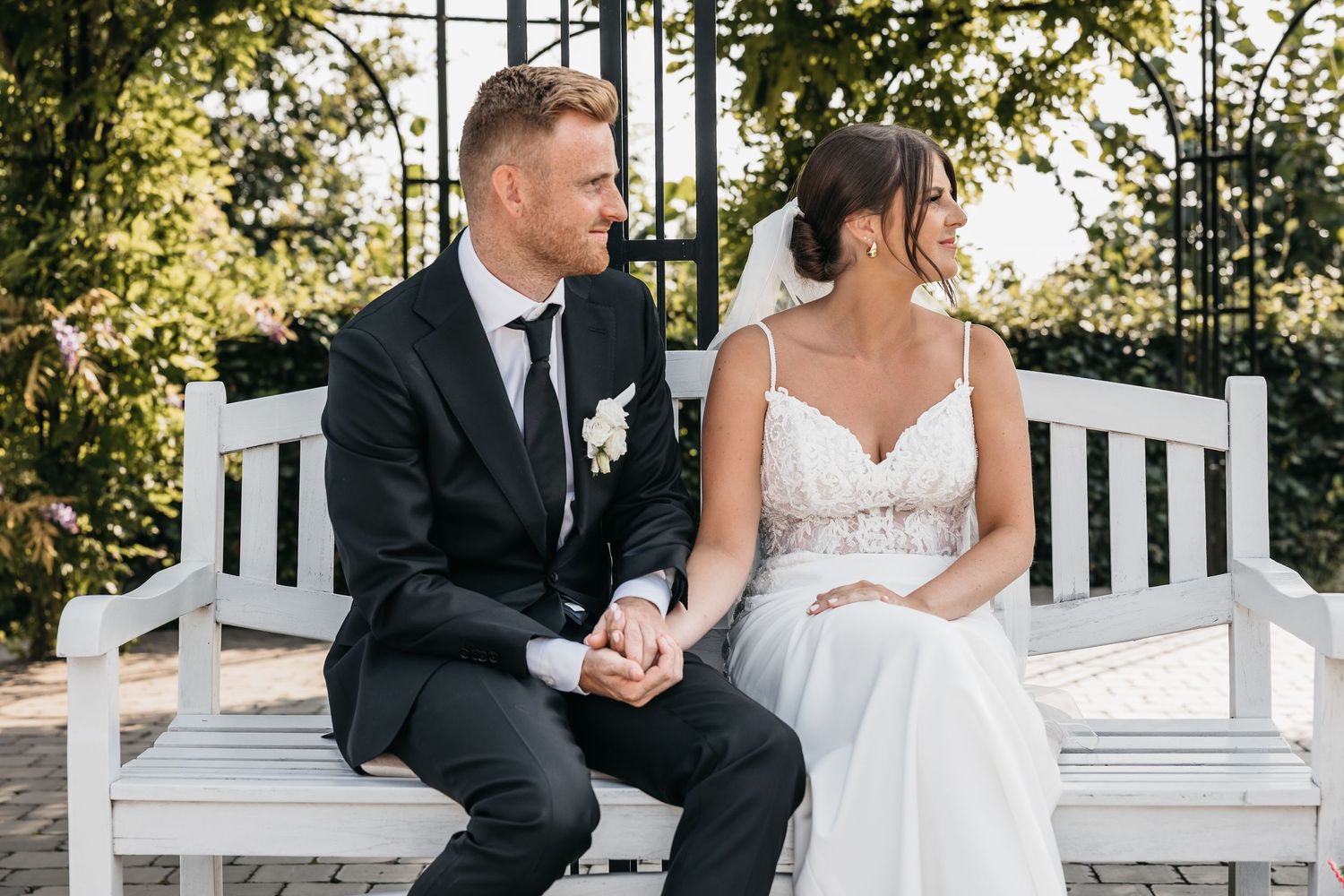 Bride and groom holding hands and gazing at each other while seated on white bench in garden.