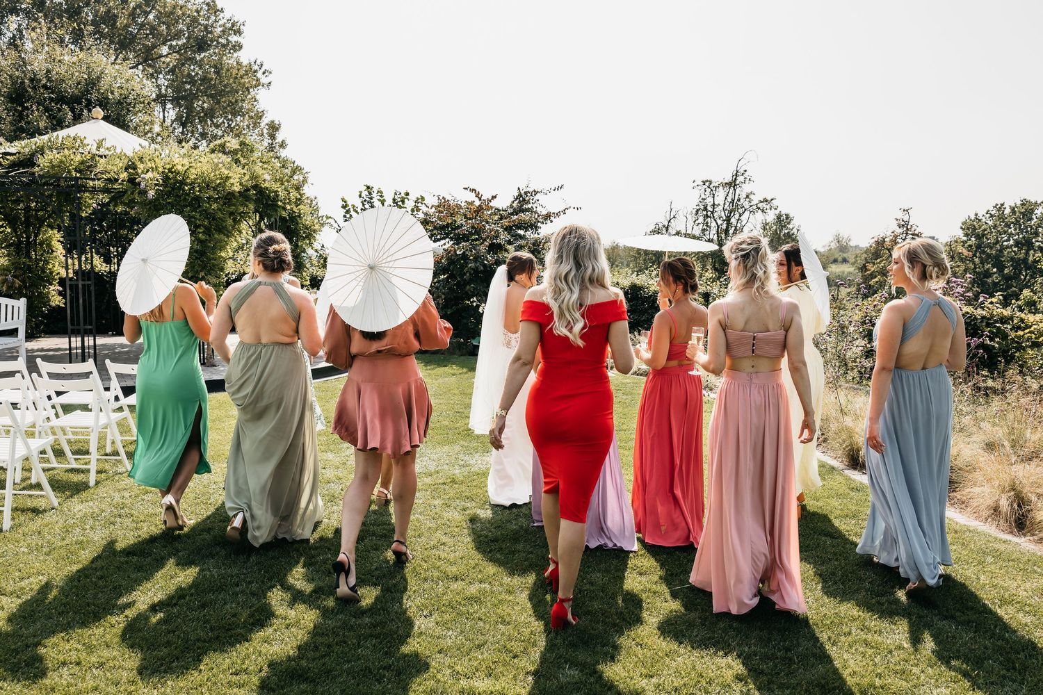 Back view of bridesmaids in multicolored dresses carrying white parasols walking toward wedding ceremony.