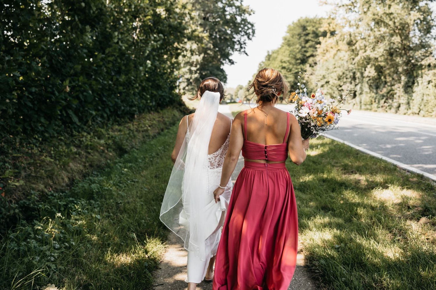 Bride in white gown and bridesmaid in red dress walk along tree-lined path outdoors.