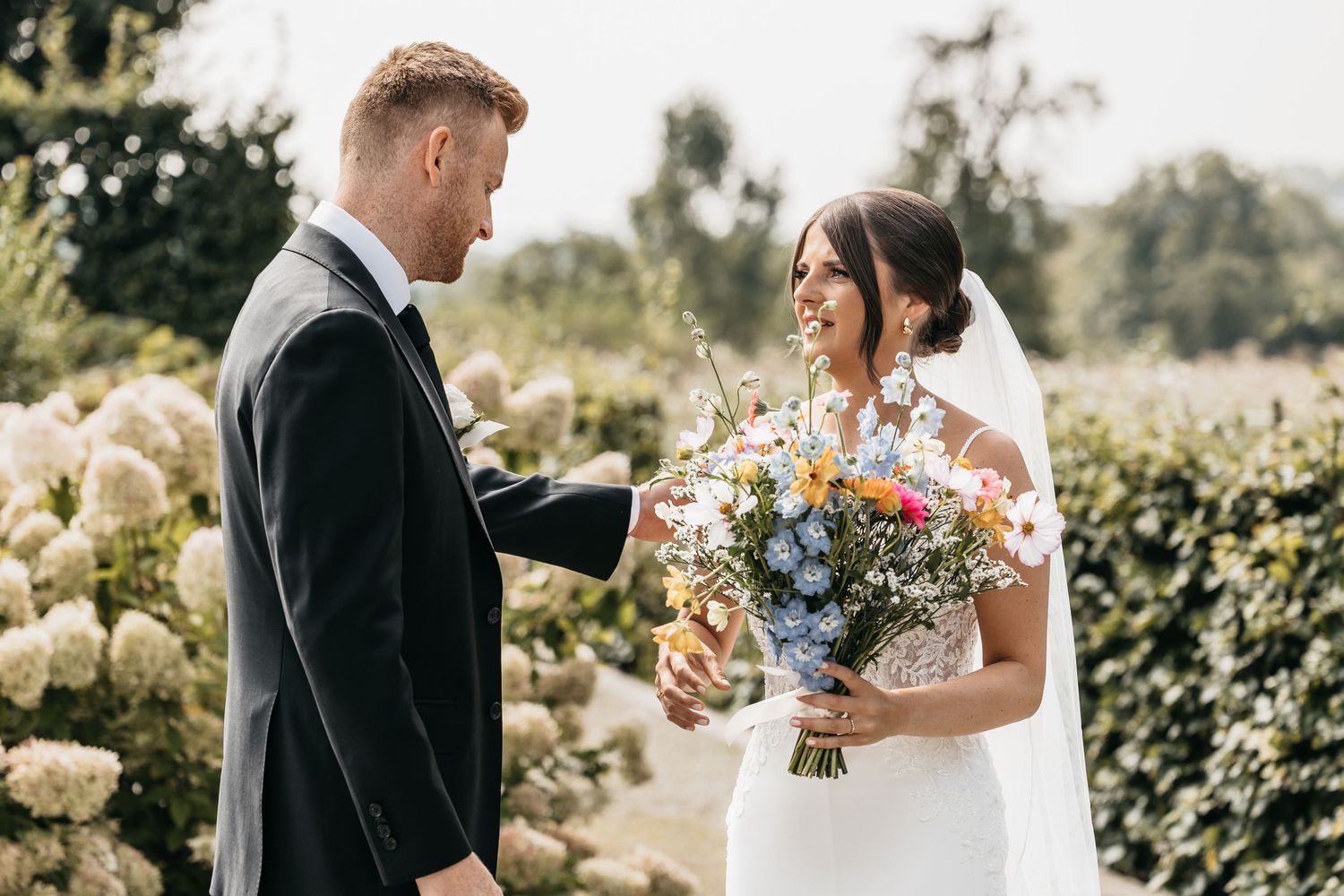 Smiling bride in lace gown holds colorful wildflower bouquet while groom in black suit stands beside her.