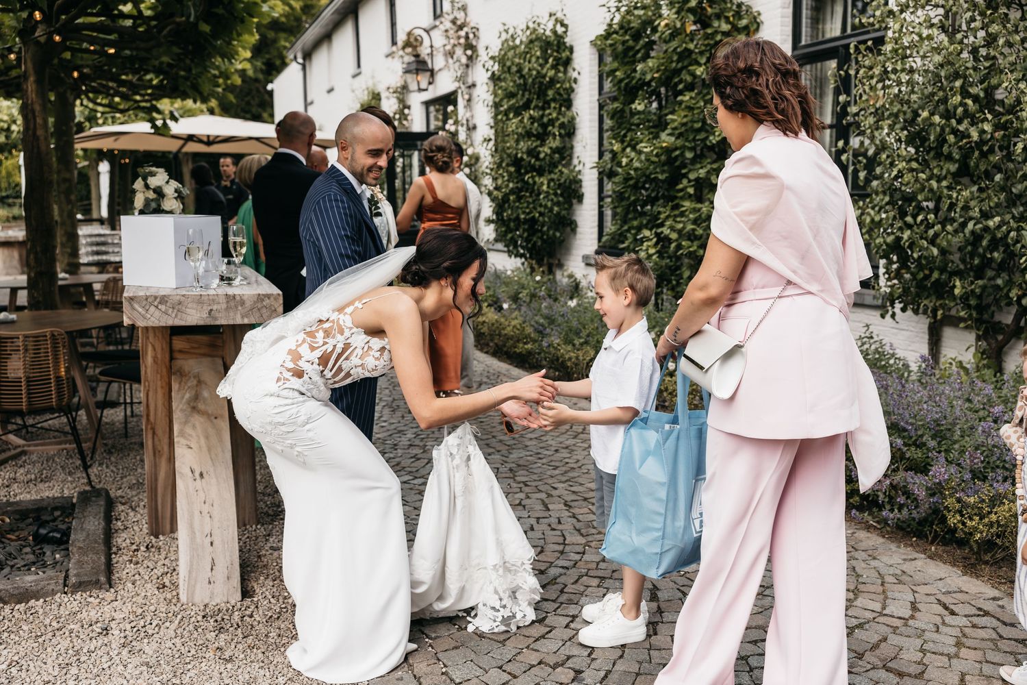 Bride bending down to greet a young flower girl at an outdoor wedding reception on a cobblestone patio.