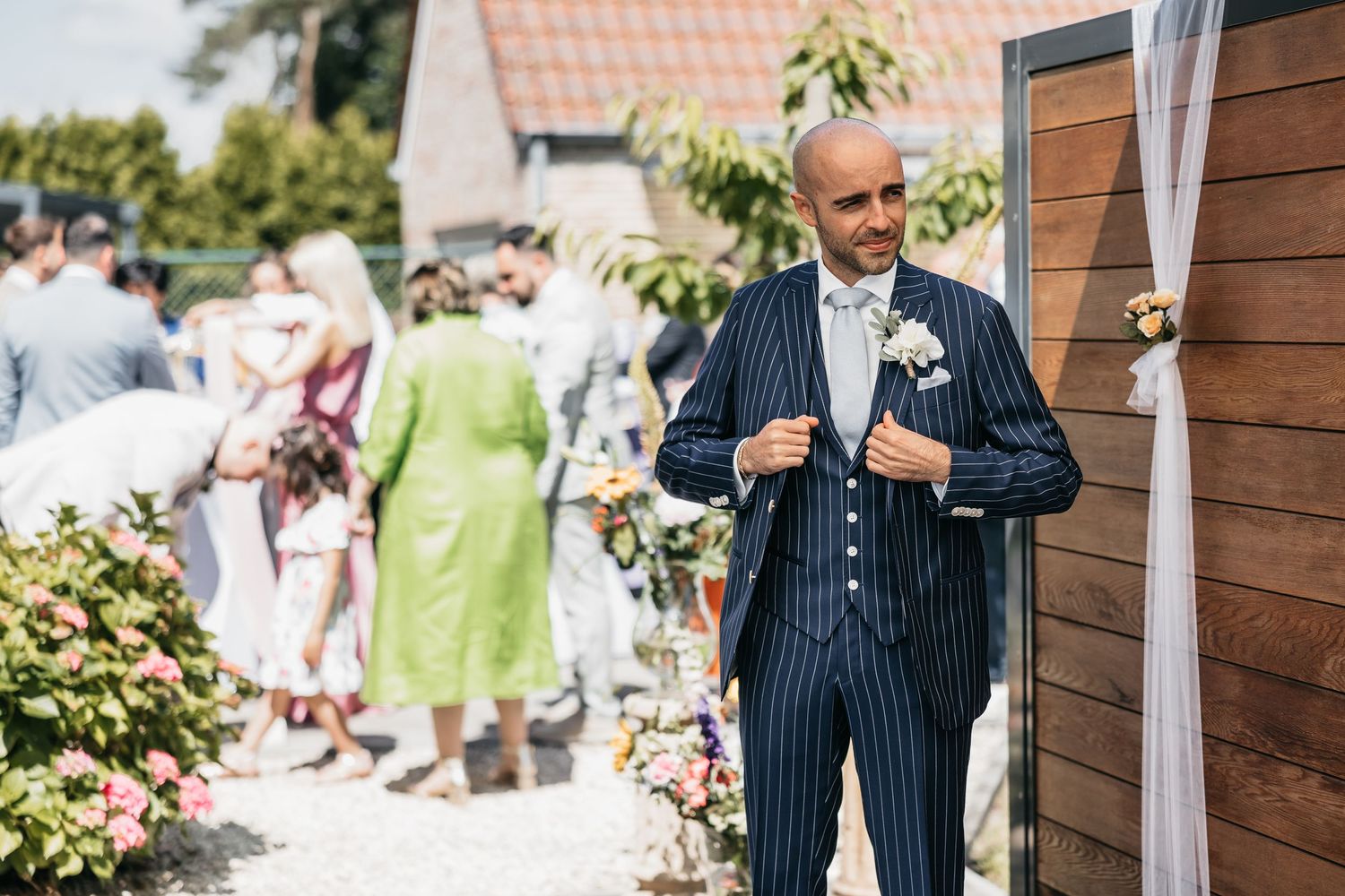 Elegant bald groom in navy pinstripe suit with white boutonniere at sunny outdoor wedding.