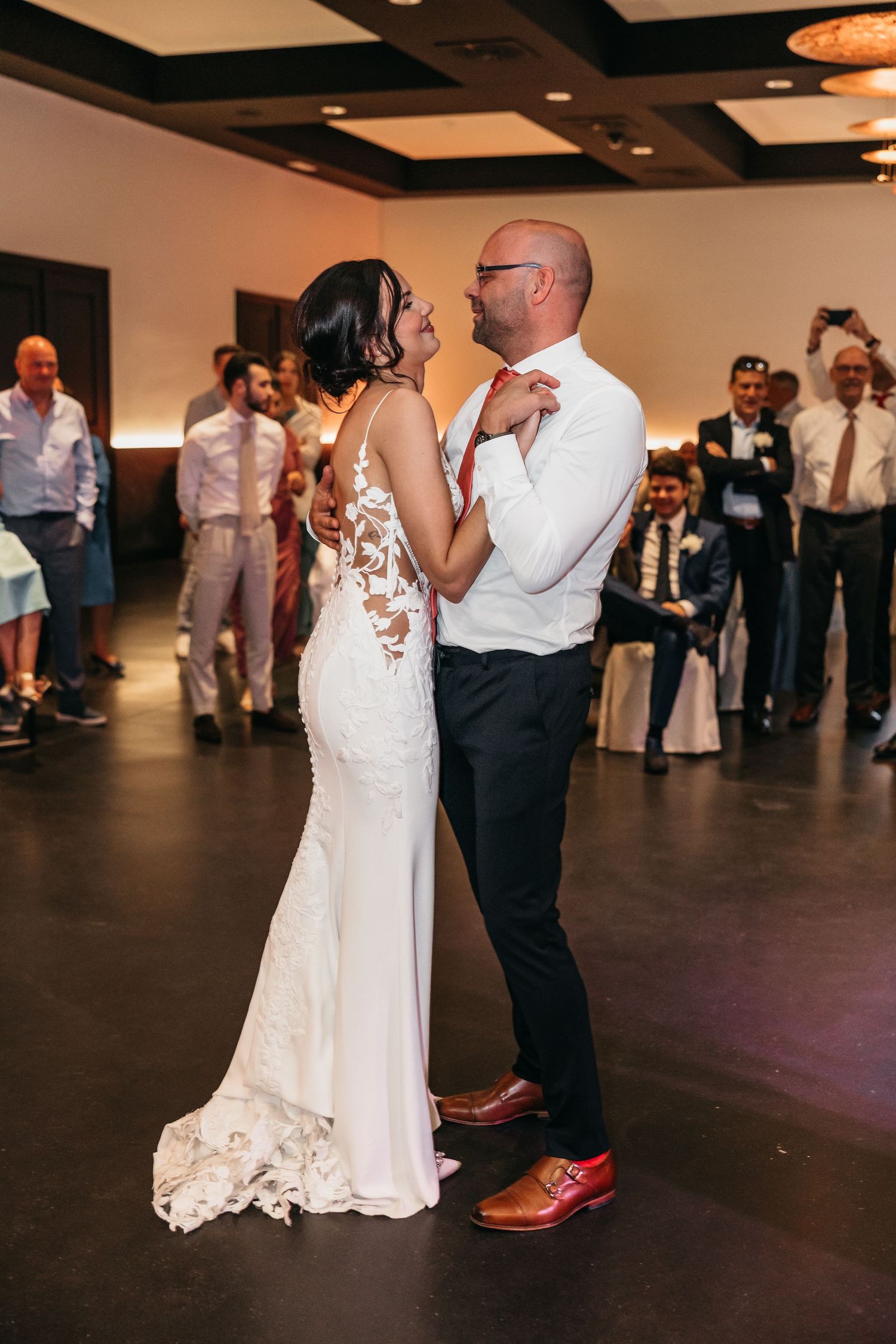 Bride in lace mermaid gown and groom in white shirt dancing at wedding reception surrounded by cheering guests.