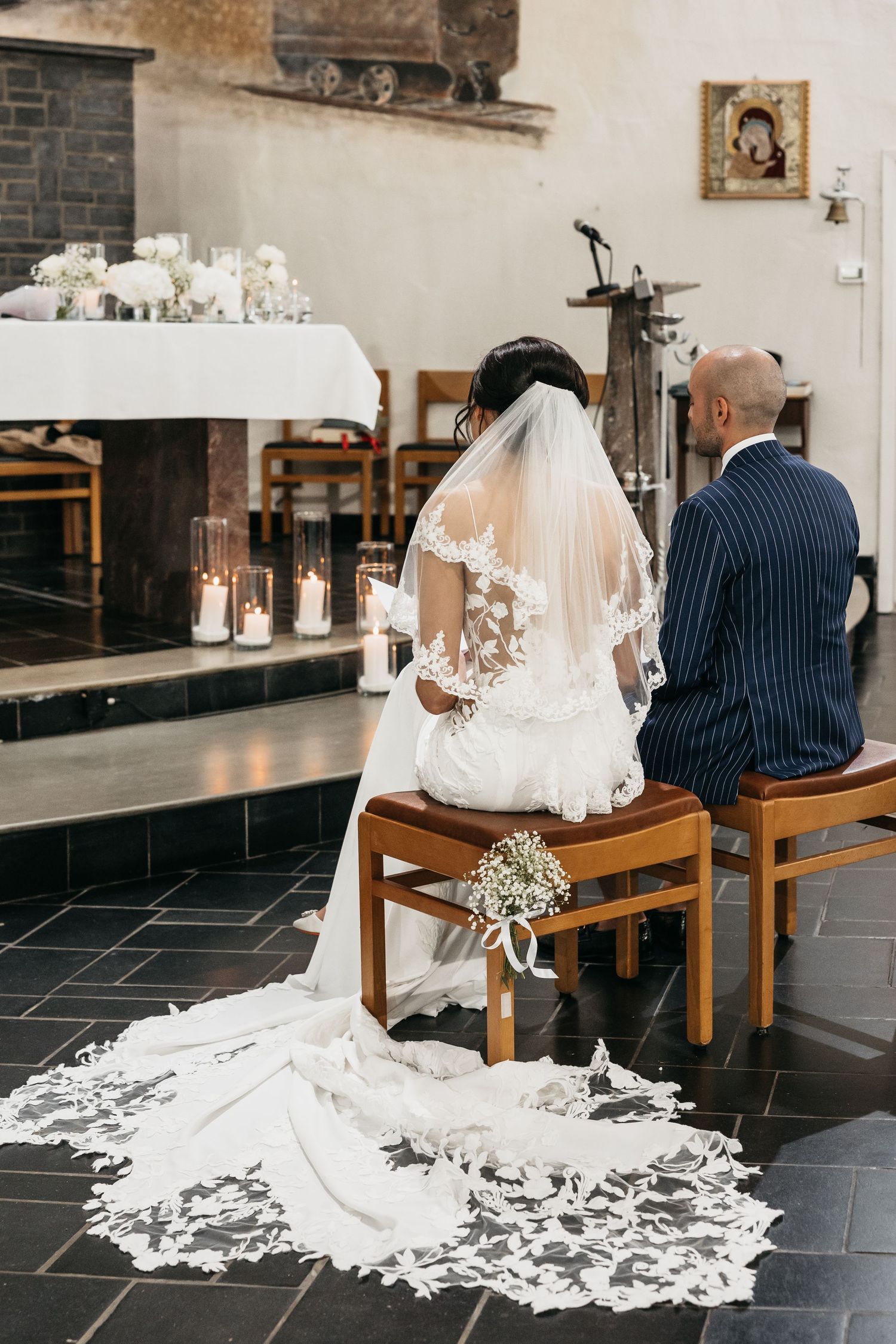 Bride in lace veil and groom sit before candlelit altar in church.