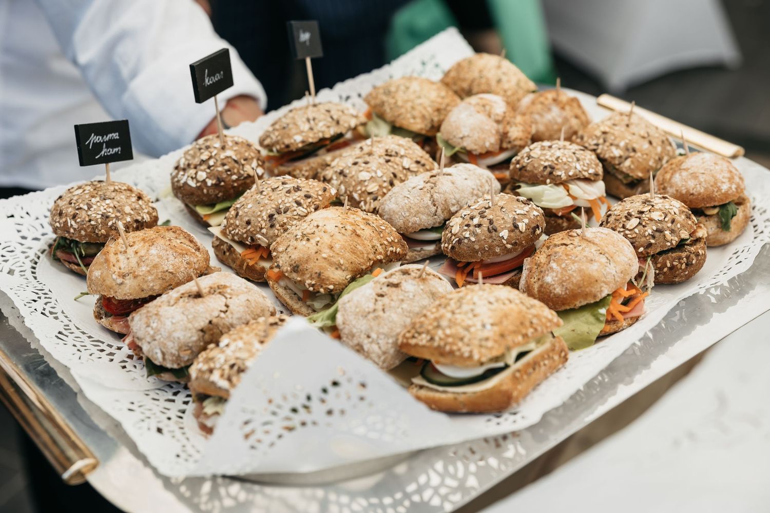 Server holding a tray of assorted mini sandwiches on seeded rolls at a catered event.