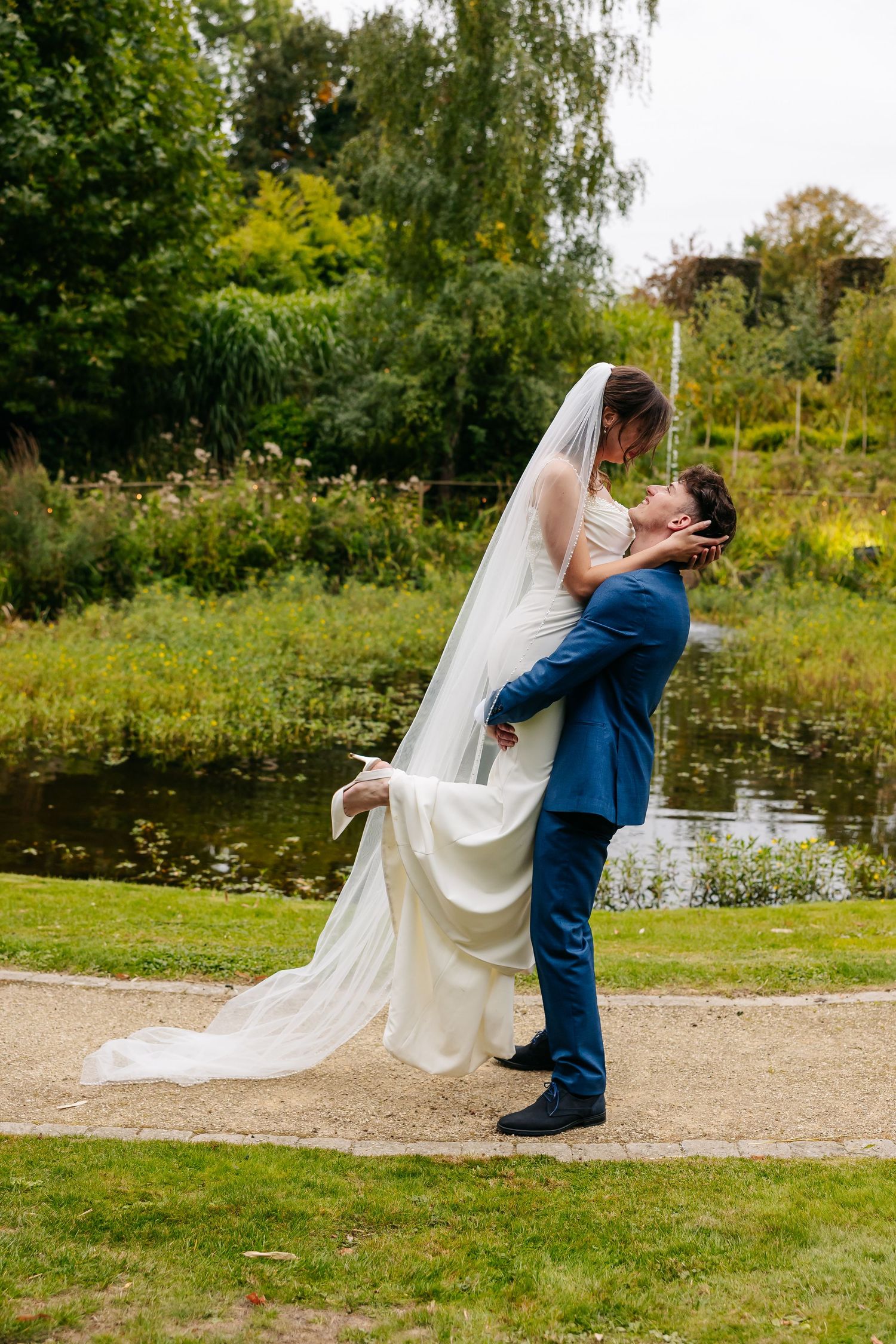 Groom lifting bride in white gown and veil for a kiss beside a tranquil garden pond.