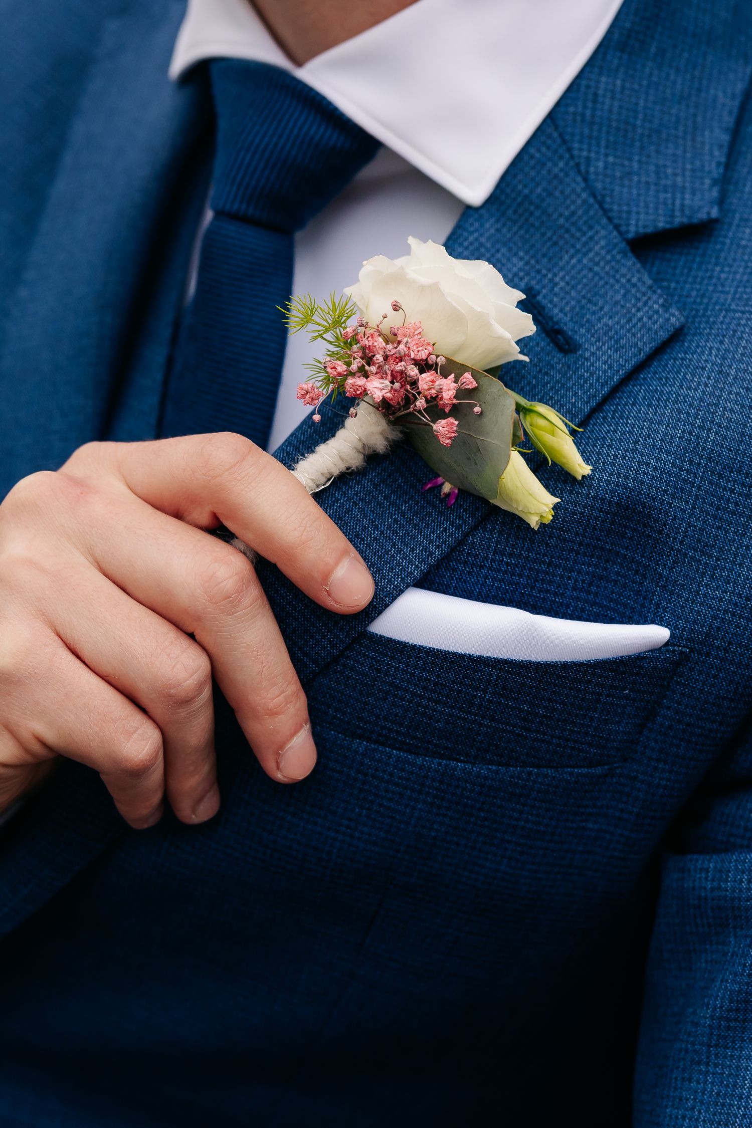 Close-up of groom in blue suit adjusting white rose and pink flower boutonniere on lapel.