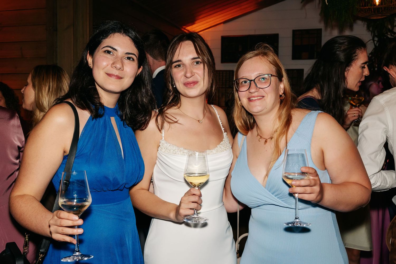 Three women in elegant dresses smile holding wine at a wedding.