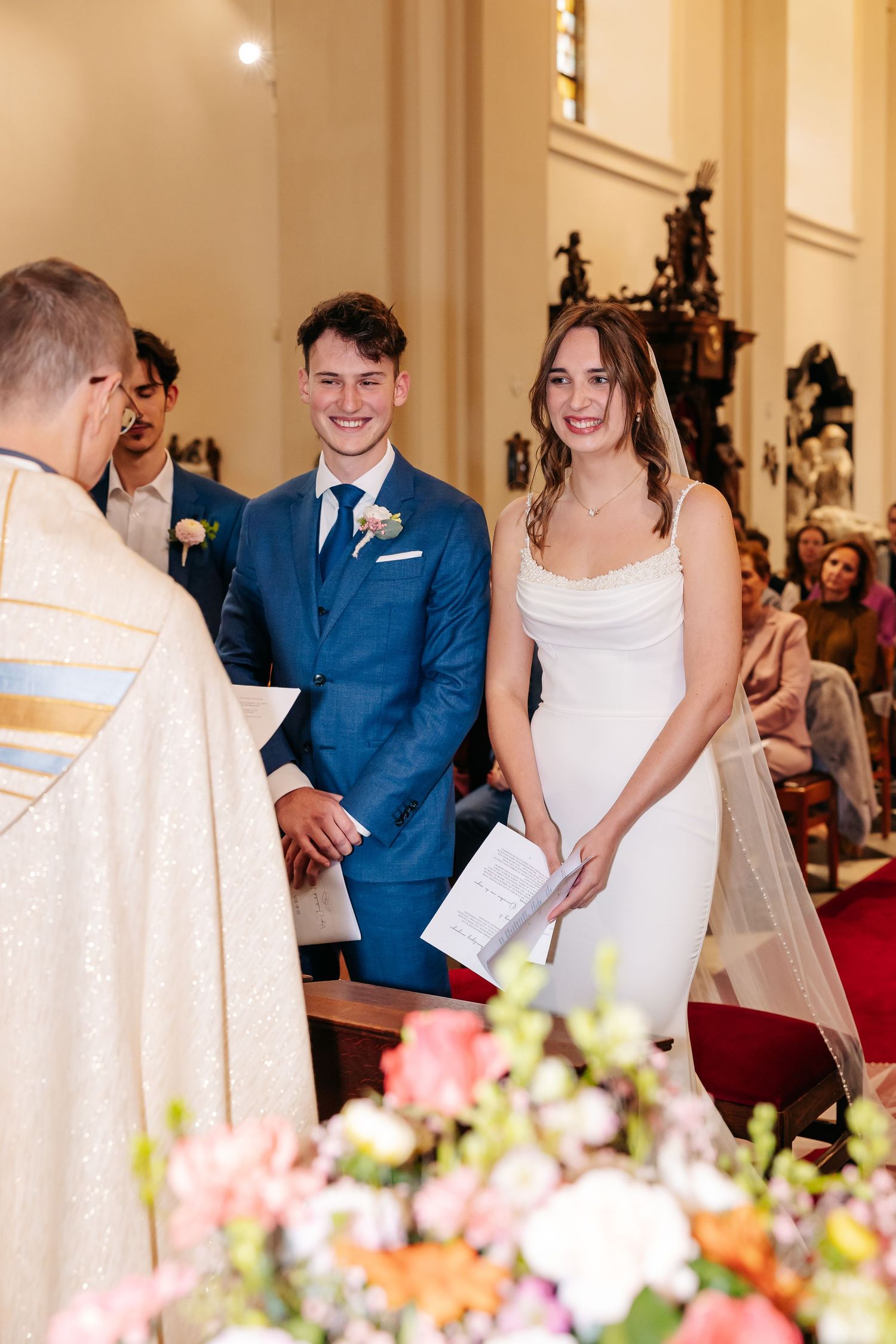 Smiling bride in white gown and groom in blue suit stand before priest during church wedding ceremony.