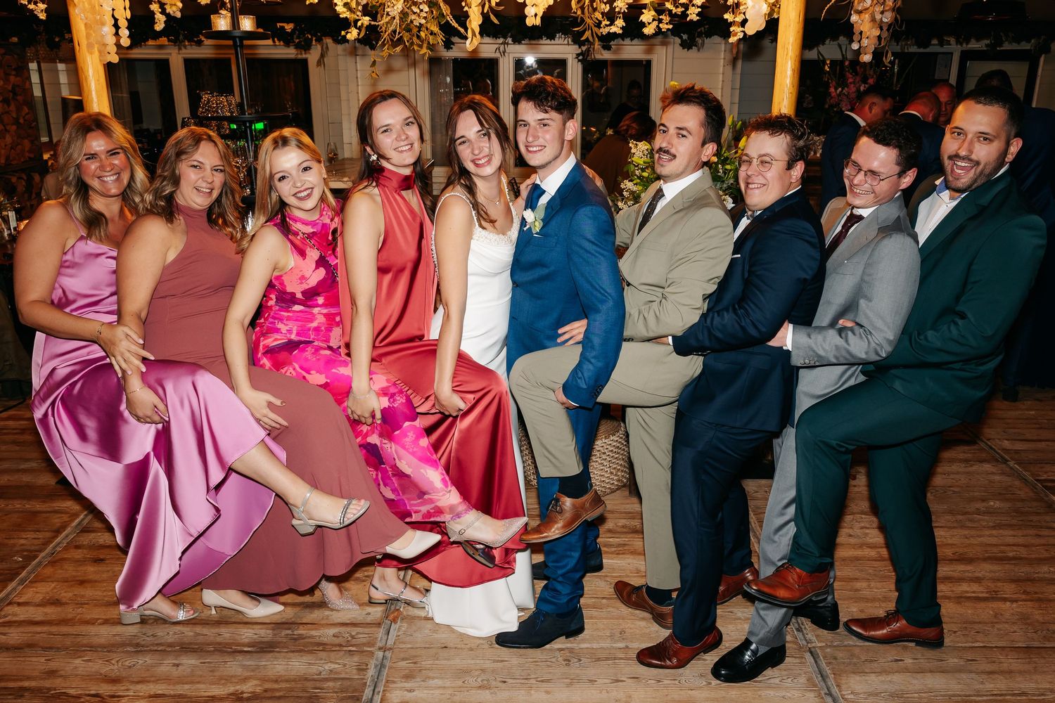 Wedding party group photo with bridesmaids in pink satin gowns and groomsmen in suits posing together.
