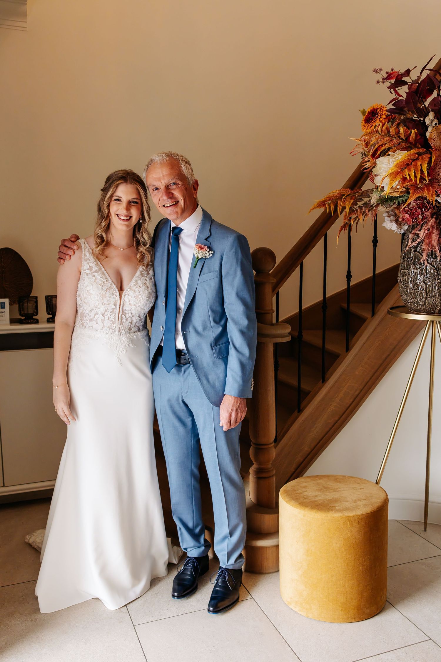 Smiling bride and older man in blue suit pose together by staircase with autumn floral arrangement.