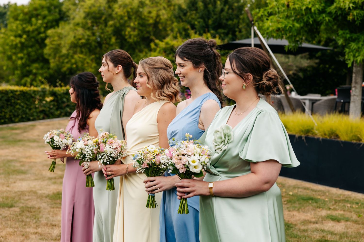 Five bridesmaids in pastel multicolored dresses holding floral bouquets at outdoor wedding.