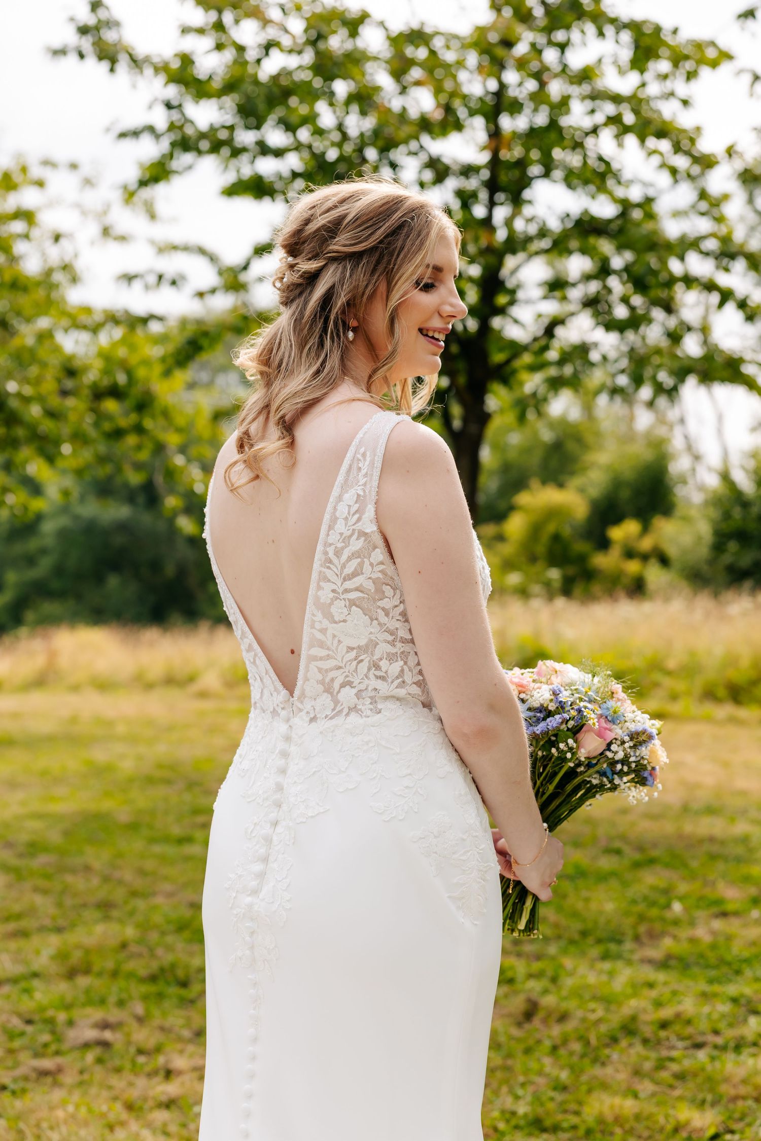 Smiling bride in a backless lace wedding gown holding a colorful bouquet outdoors.