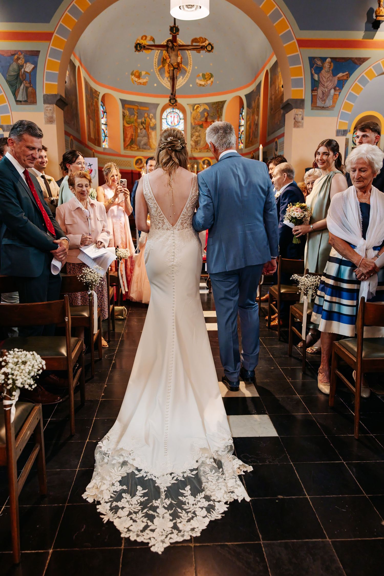 Bride in lace backless gown walks down church aisle escorted by father toward altar.
