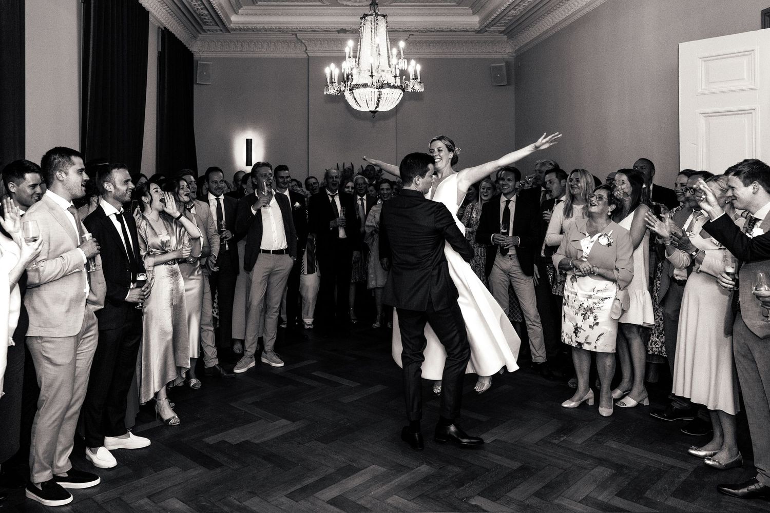 Black and white photo of bride and groom dancing energetically surrounded by applauding guests.