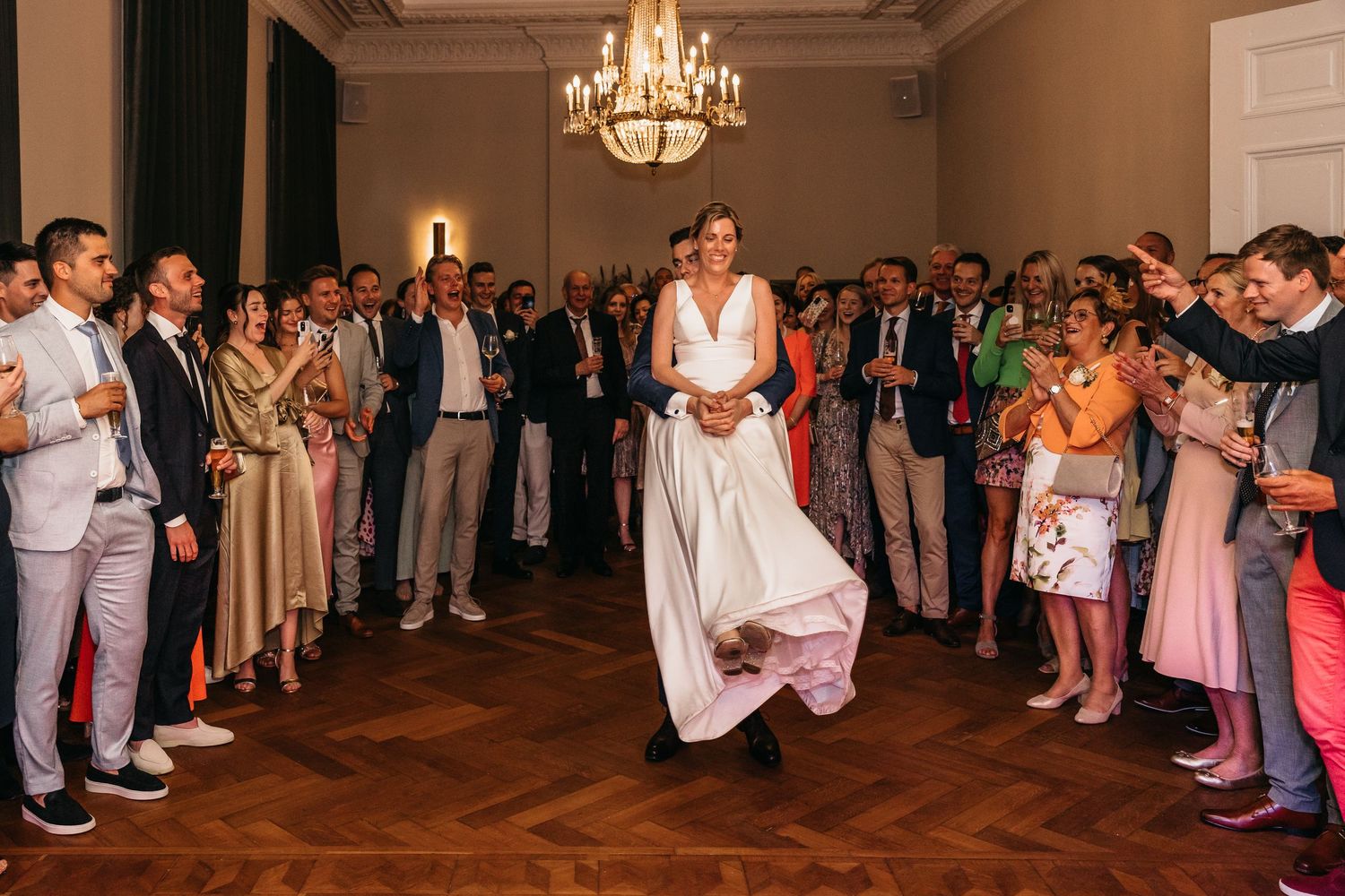 Bride in white gown dancing joyfully surrounded by cheering guests at reception.
