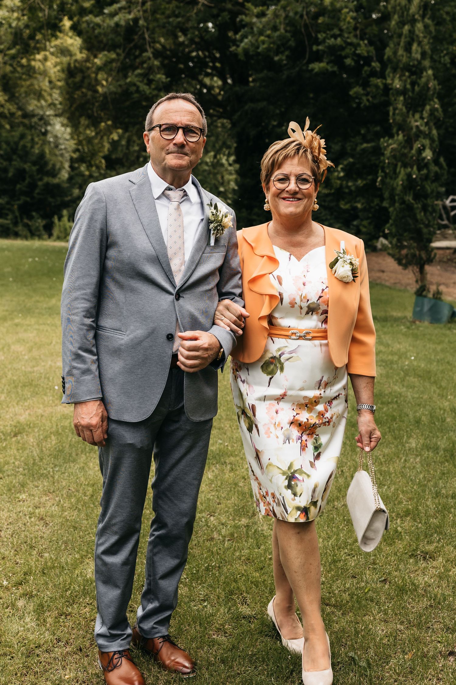 Smiling couple in elegant wedding guest attire pose together on green lawn at outdoor wedding.