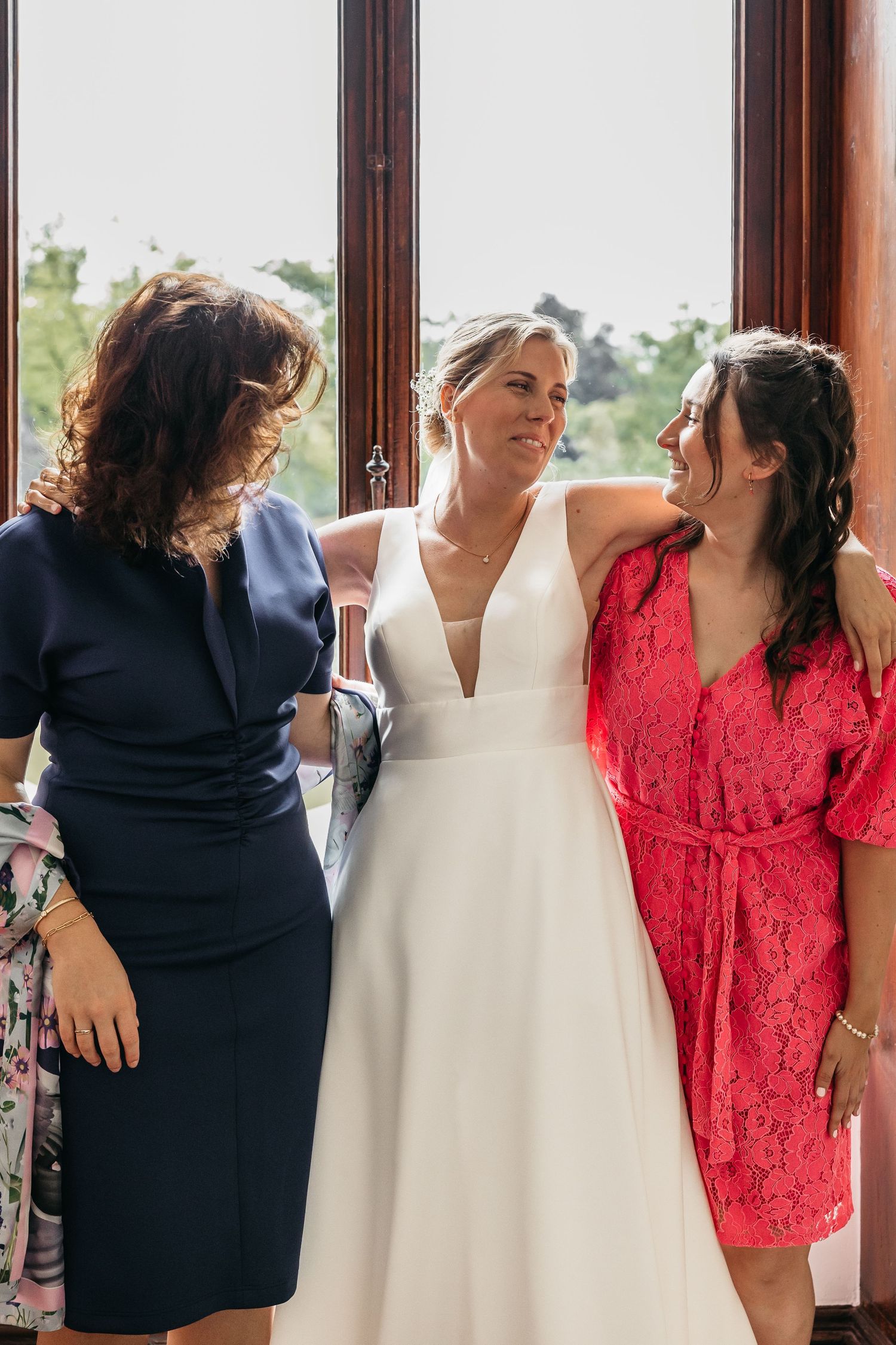 Bride in white wedding dress standing between two women in navy and red outfits indoors.