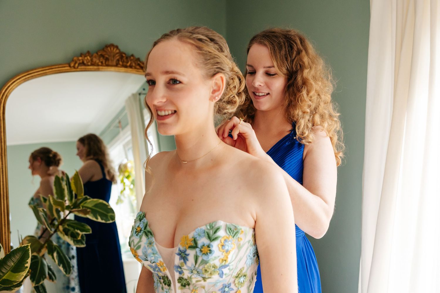 Curly-haired bridesmaid in blue dress adjusting bride's earring near gold ornate mirror.