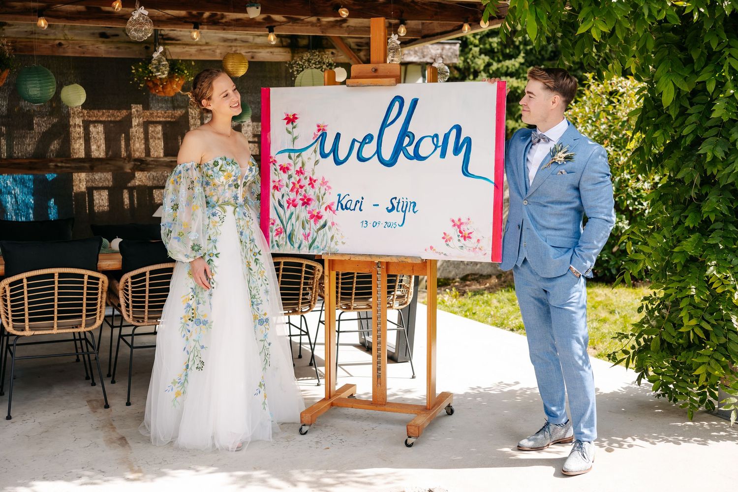 Bride and groom stand beside a colorful welcome sign reading Welkom at their outdoor wedding venue.
