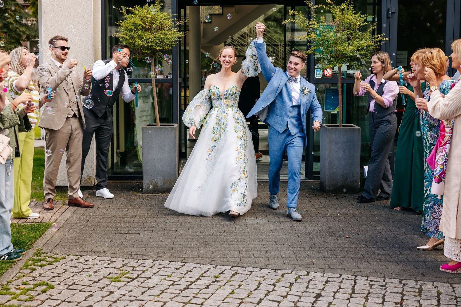 Bride in floral gown and groom in blue suit exit venue as guests celebrate their wedding.