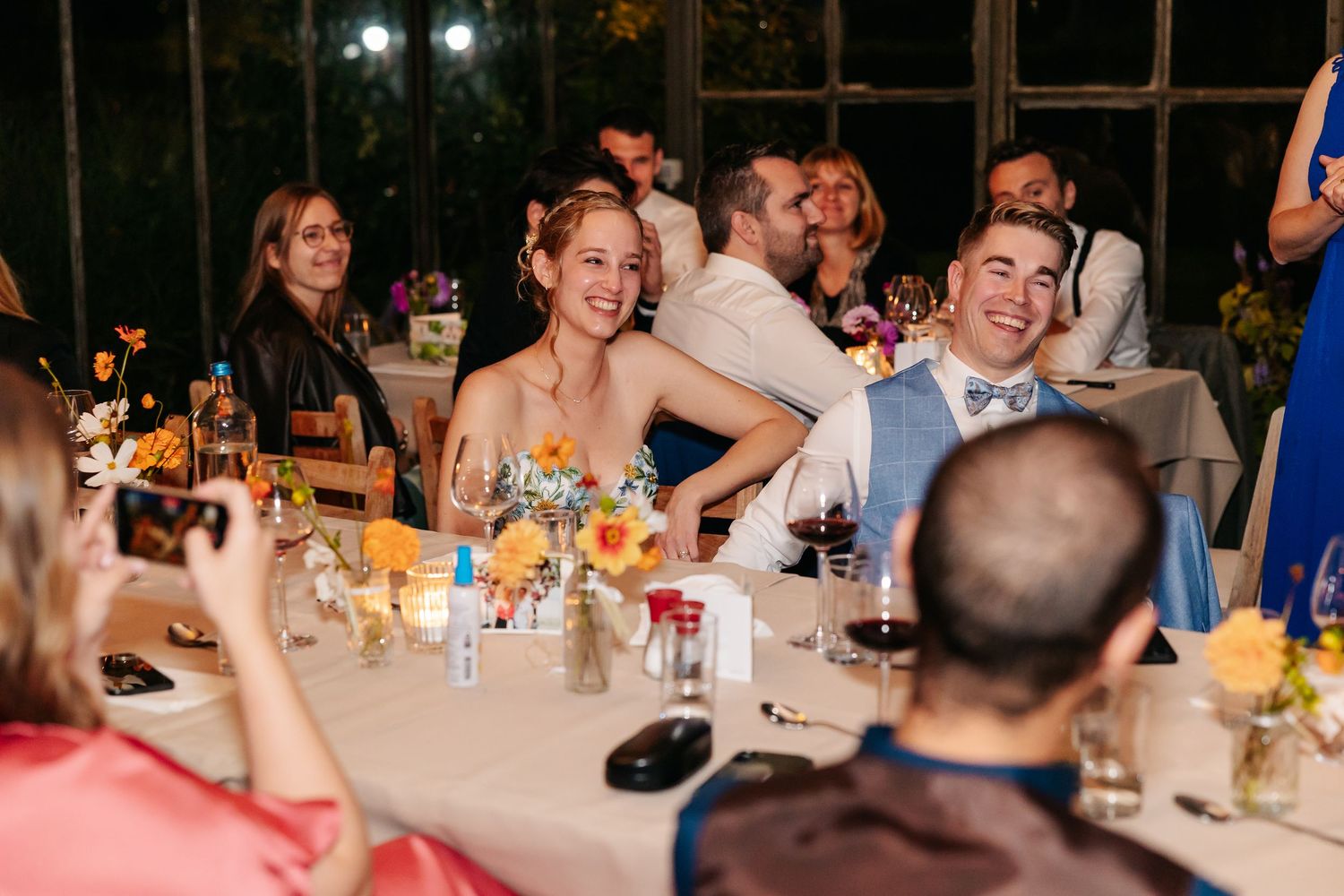 Bride and groom laugh joyfully at their wedding reception table surrounded by colorful floral centerpieces.