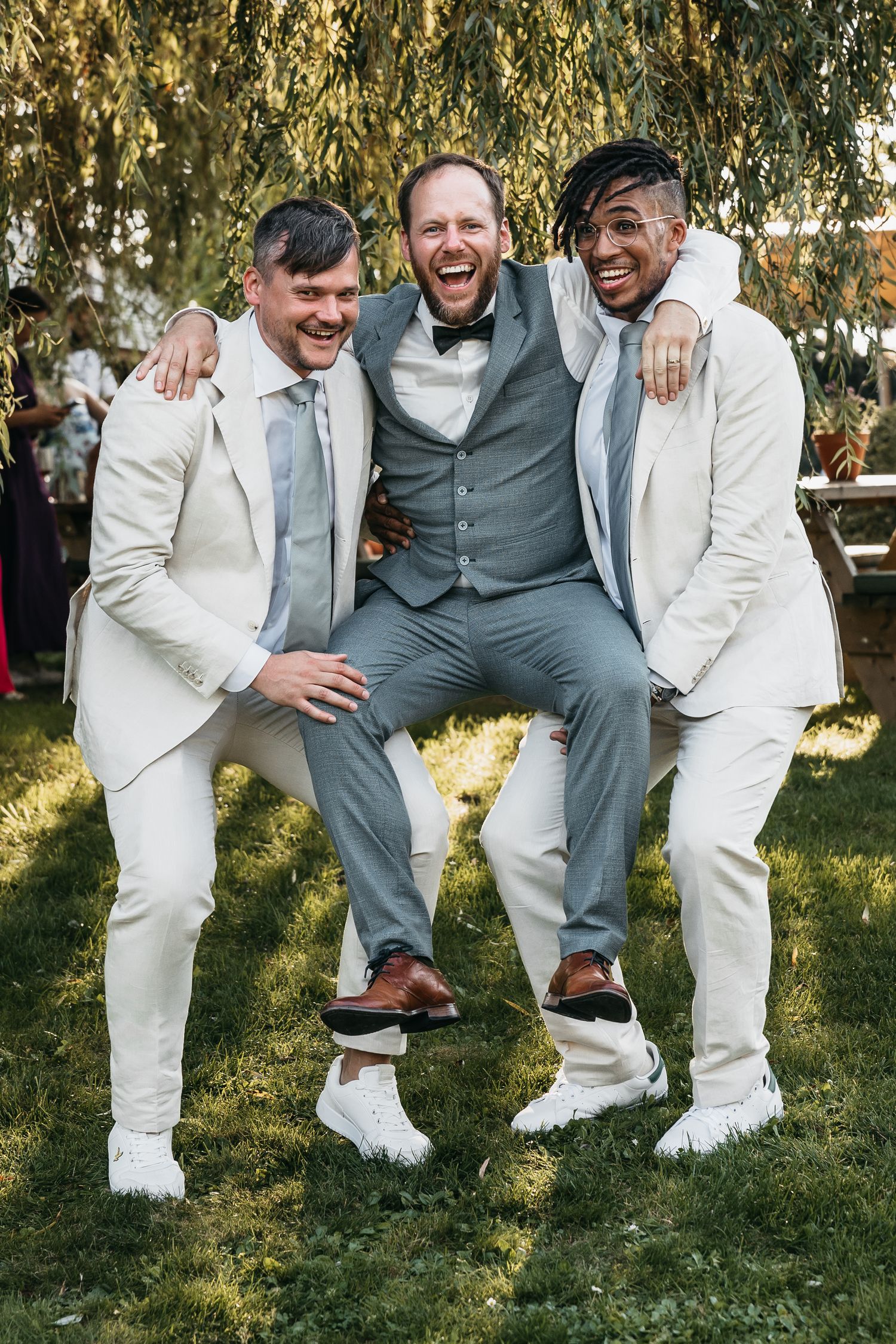 Two groomsmen in cream suits playfully lifting the laughing groom in a gray vest outdoors.