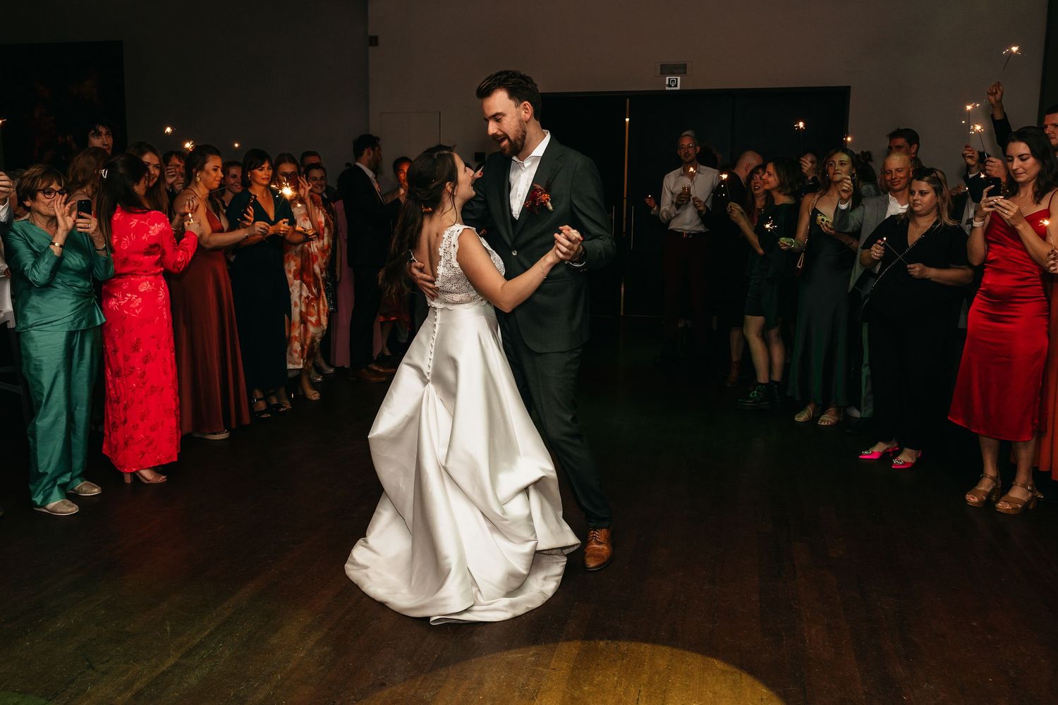 Bride and groom share their first dance surrounded by guests holding sparklers at wedding reception.