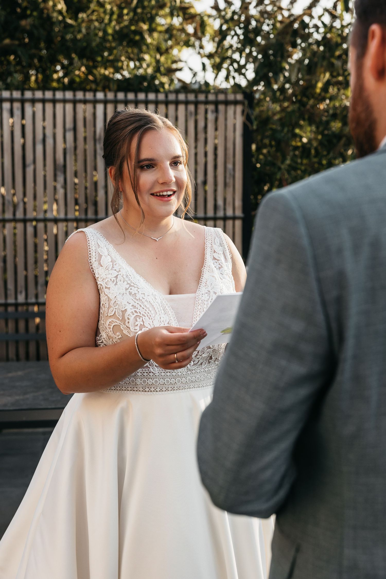 Radiant bride in lace wedding gown smiles while reading heartfelt vows to groom during private outdoor ceremony.