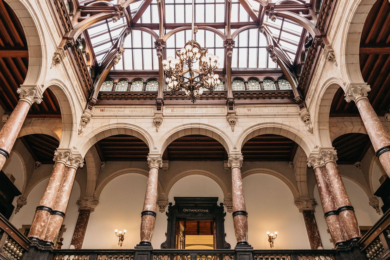 Grand historic building interior with arched galleries, marble columns, and ornate glass ceiling chandelier.