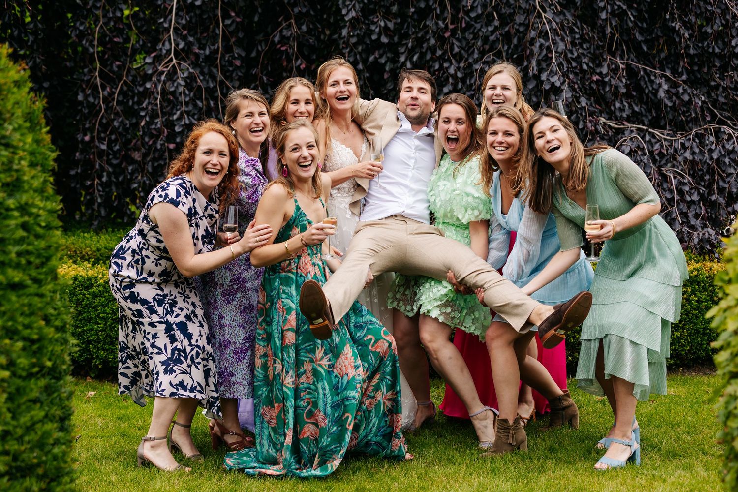 Groom being lifted and surrounded by a group of laughing bridesmaids and female guests outdoors.