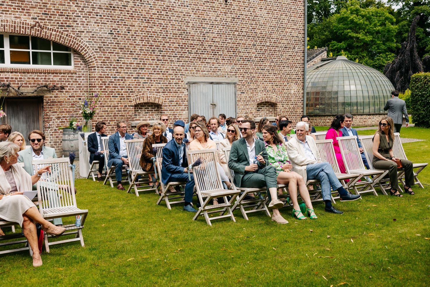 Wedding guests seated on wooden chairs on a sunny lawn during an outdoor ceremony.