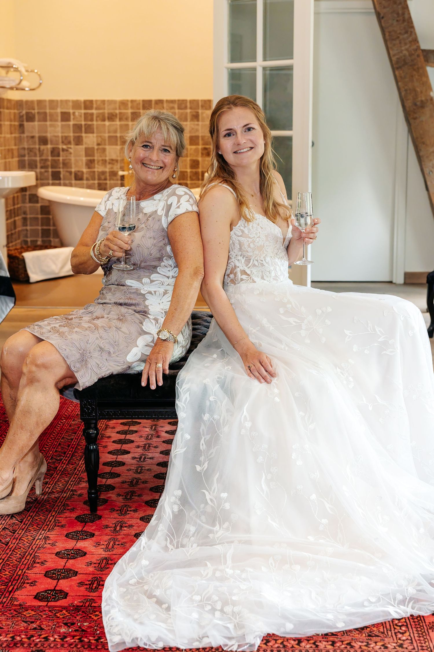 Bride in white lace gown and older woman in silver dress toasting champagne glasses indoors.