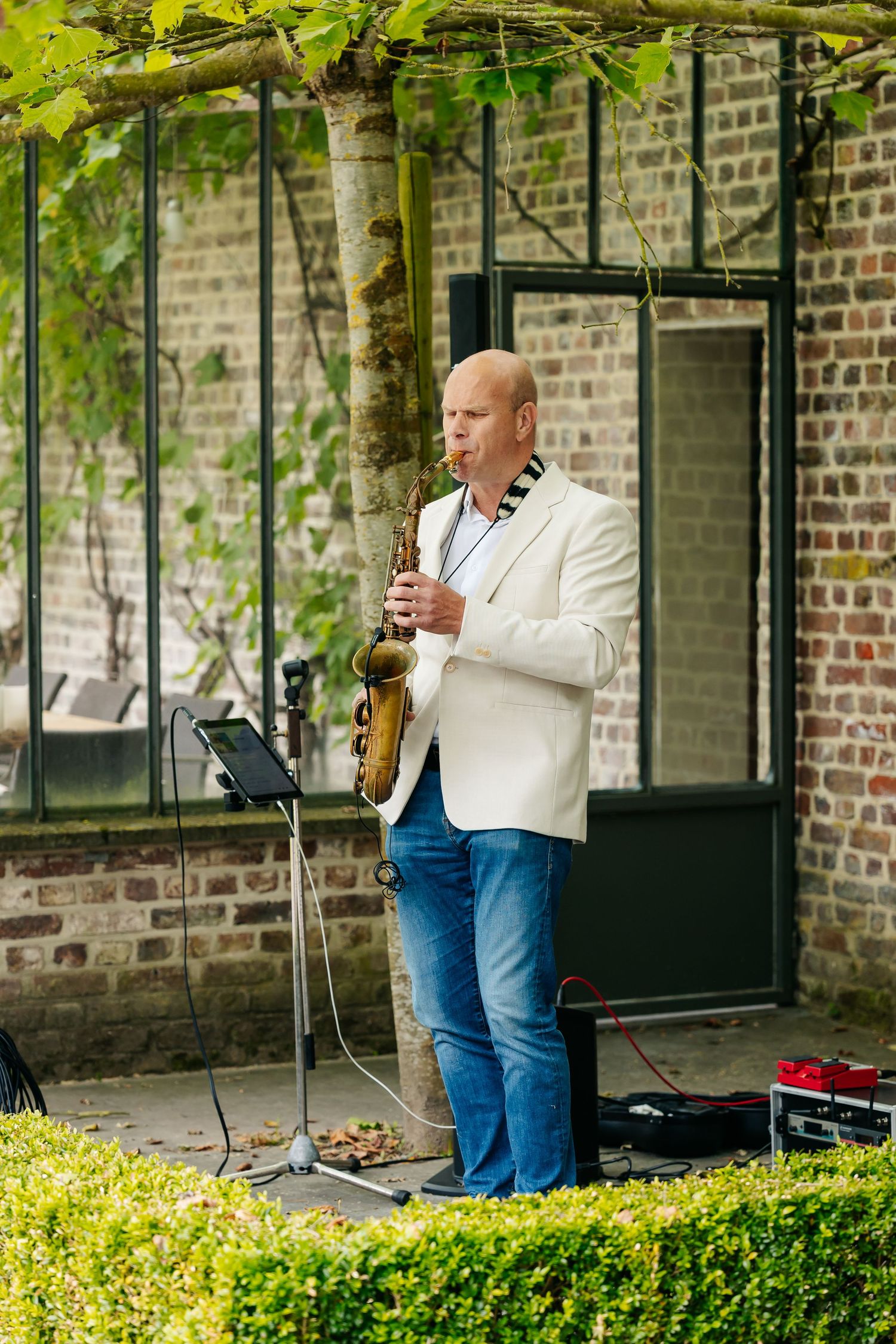 Bald saxophonist in white jacket performs outdoors at wedding reception near brick wall.