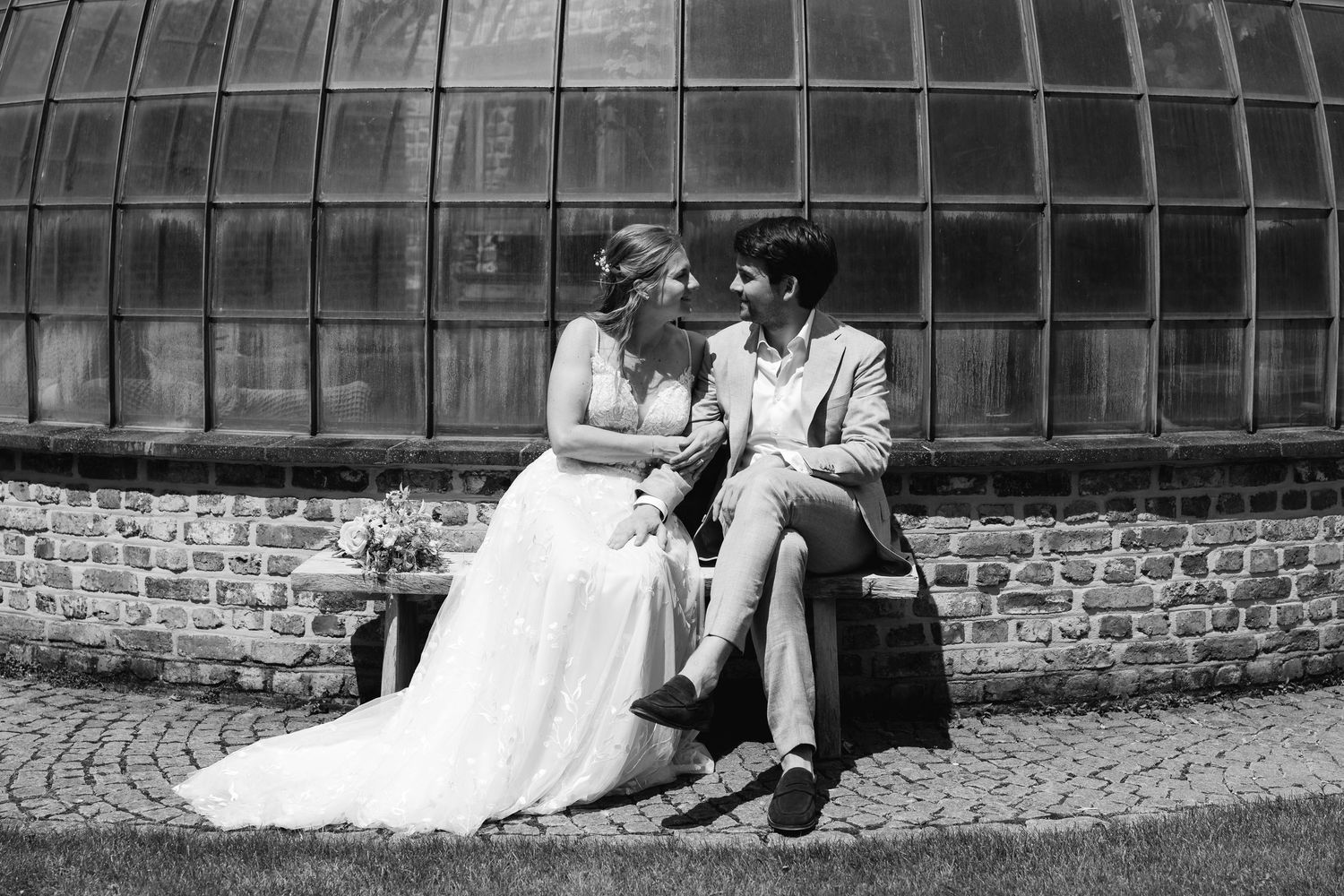 Black and white photo of a bride and groom sitting together on a bench outside a greenhouse.